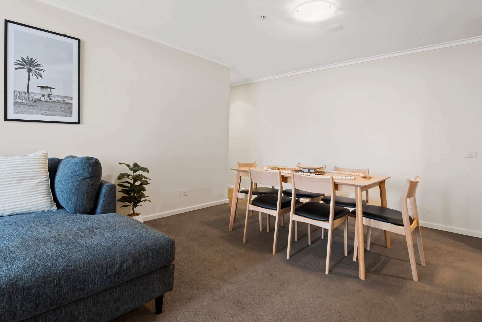 Una habitación con un sofá azul, una mesa de comedor con sillas, un cuadro en la pared y una planta.