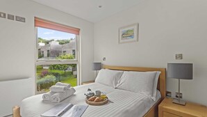 A hotel room with a bed, a bedside table, a lamp, a window with a view of buildings and greenery, and a framed picture on the wall.