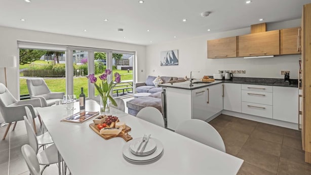 A modern kitchen with white cabinets, a dining area with a white table set for a meal, and a view of a garden through large sliding doors.