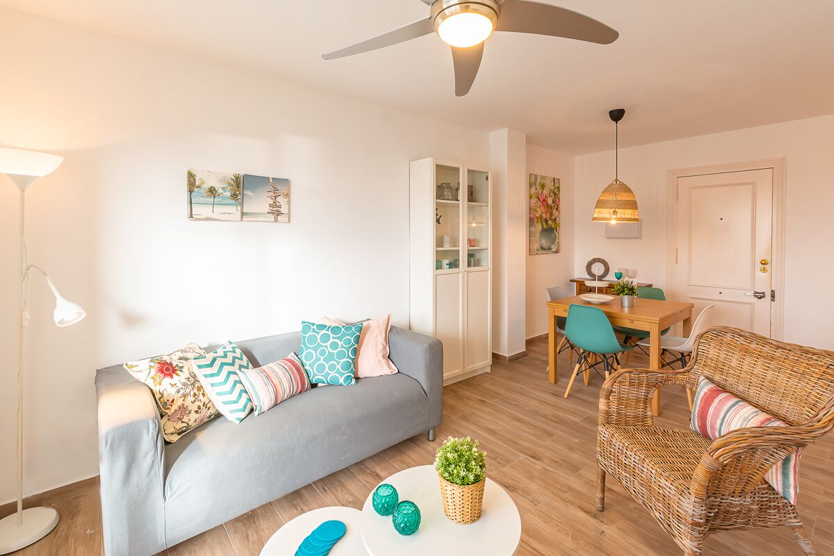 Een moderne woonkamer met een grijze bank, een rieten fauteuil en een eethoek met een tafel en stoelen.