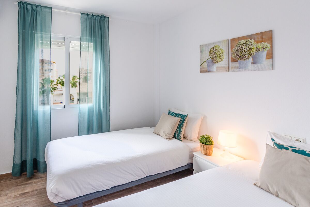 A hotel room with two beds, a window with teal curtains, a bedside table with a lamp, and a wall-mounted plant display.