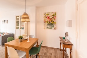 A dining area with a wooden table, green and white chairs, a hanging light fixture, and a wall-mounted painting of flowers.