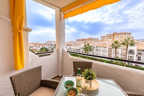 A balcony with wicker furniture, a table with a bottle and glass, and a view of buildings and palm trees.
