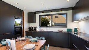 A modern kitchen with dark cabinetry, a wooden dining table set for six, and a large window providing natural light. - Villa Lion | Private Spa, Pool/Jacuzzi, Terrace, Arcade (Ittenheim)