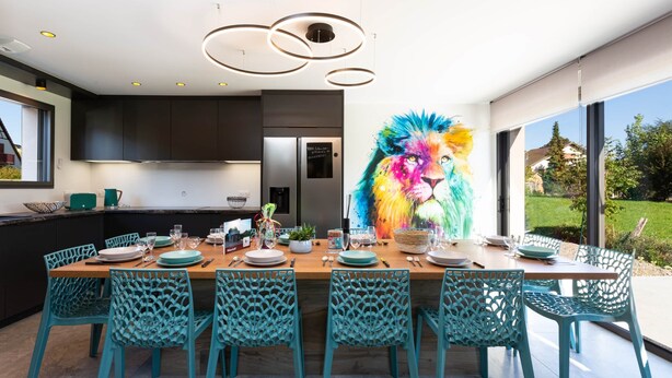 A modern kitchen with a large window, a colorful lion mural, and teal chairs around a wooden dining table. - Villa Lion | Private Spa, Pool/Jacuzzi, Terrace, Arcade (Ittenheim)