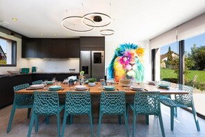 A modern kitchen with a large window, a colorful lion mural, and teal chairs around a wooden dining table. - Villa Lion | Private Spa, Pool/Jacuzzi, Terrace, Arcade (Ittenheim)