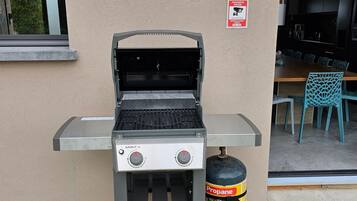 An outdoor grill with propane tank, a window, and a door leading to an indoor area with chairs.