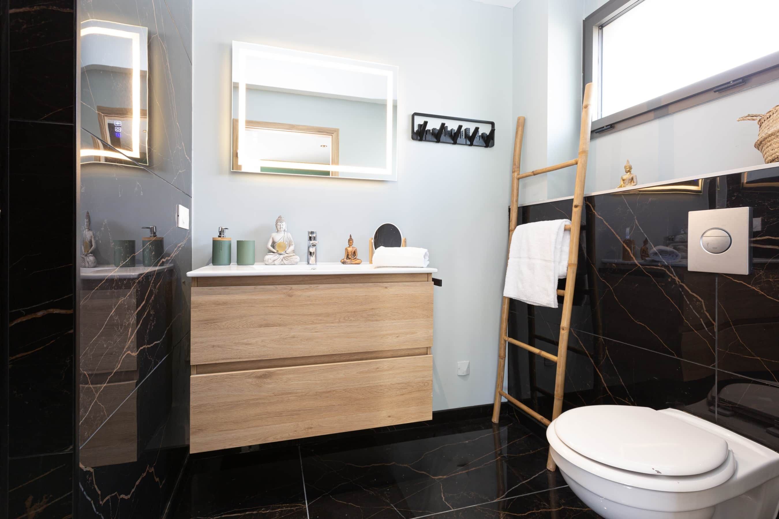 Et moderne badeværelse med marmorgulv, en trækommode, toilet og et spejl med indbygget lys.