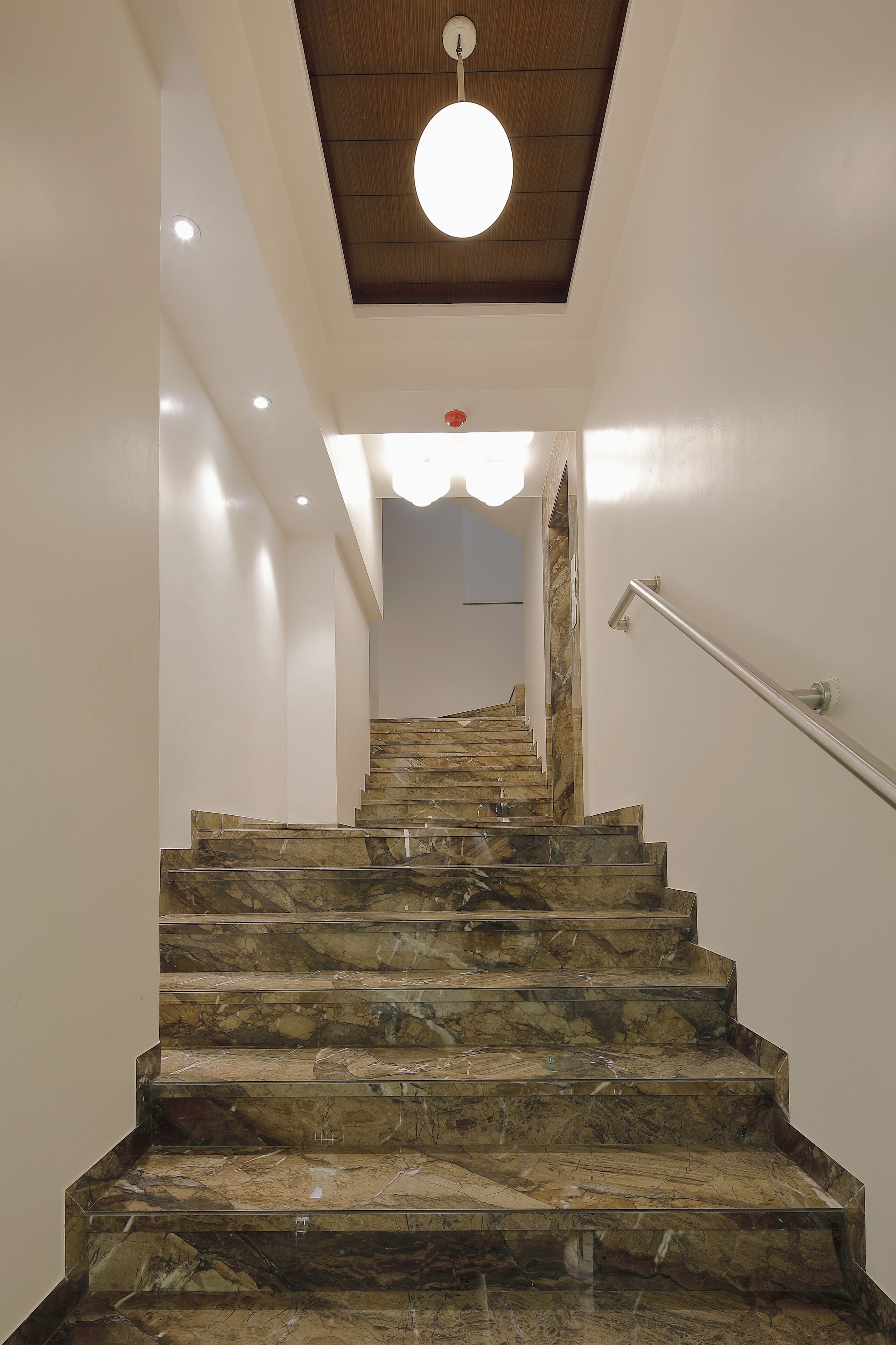 Un escalier avec des marches en marbre et une rampe, menant à un palier éclairé par un luminaire au plafond.