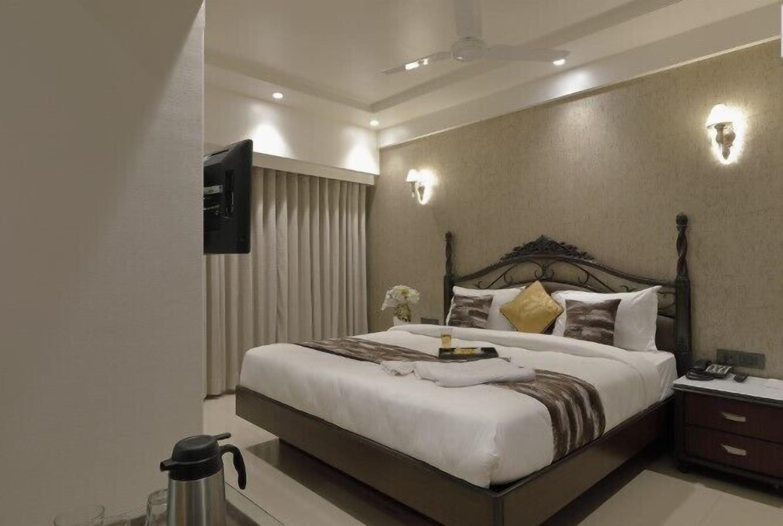 Ein modernes Hotelzimmer mit einem großen Bett, einem Nachttisch mit Lampe, einem an der Wand montierten Fernseher und einer Kaffeemaschine auf dem Boden.