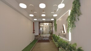 Zona de recepción de hotel con un cartel que dice 'WELCOME', un pasillo con plantas en macetas y una escalera que lleva a las habitaciones.