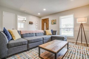A living room with a sectional sofa, a coffee table, a patterned rug, a floor lamp, and a door with a 'WELCOME' sign.
