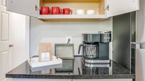 Una cocina con cafetera, un armario con tazas rojas y blancas, y una estantería con copas de vino.