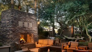 An outdoor patio area with a stone fireplace, wooden furniture, and lush greenery. - Why Wait? Reserve your Villa today! (Phoenix)