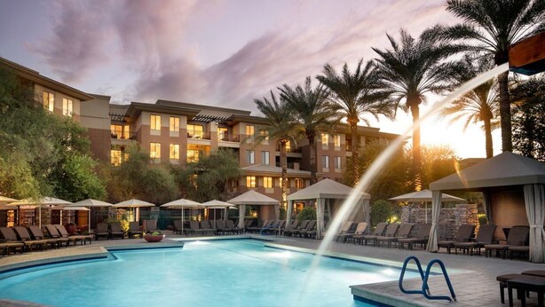 A hotel pool with lounge chairs and umbrellas, surrounded by palm trees and a multi-story building with balconies. - Why Wait? Reserve your Villa today! (Phoenix)