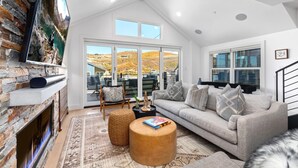 A spacious living room with a stone fireplace, a large sofa, and a view of a mountainous landscape through large windows.