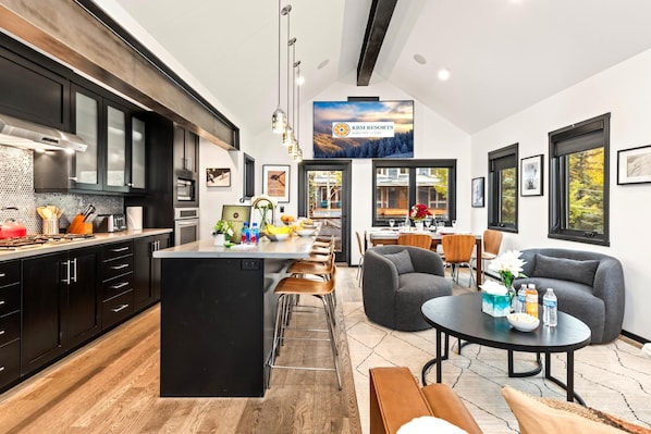 A modern kitchen with a central island, dining area with a round table, and a living room with a sofa and chairs.