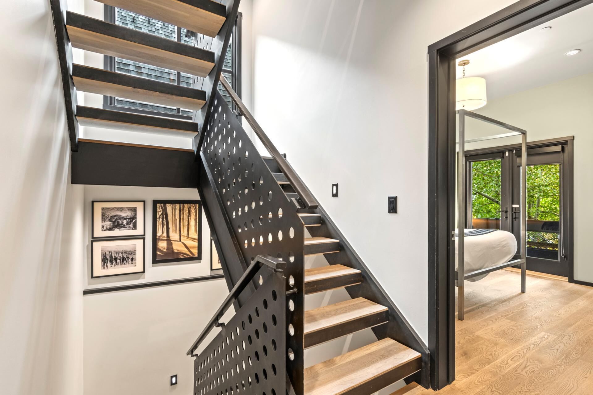 A modern staircase with a black metal railing and wooden steps, leading to a room with a bed and a view of greenery outside.