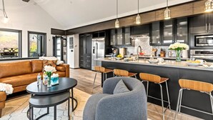 A modern kitchen with dark cabinets, stainless steel appliances, and a central island. There is a dining area with a brown leather sofa and a round table.