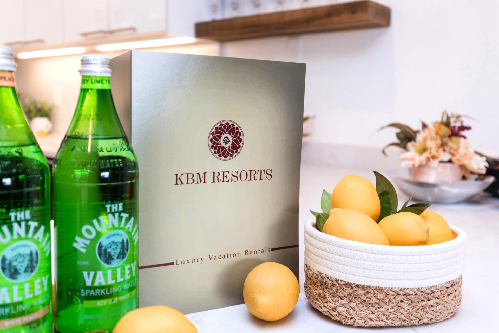 A brochure titled "KBM Resorts" with a logo, and bottles of "The Mountain Valley" sparkling water. There are lemons in a woven basket.