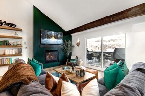 A cozy living room with a fireplace, bookshelves, and a large window offering a view of the outdoors. - Aspen Grove 3 Bedrooms AGC-8 by KBM (Park City)