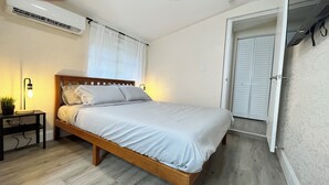 A bedroom with a wooden bed, a bedside table with a lamp, a potted plant, and a window with blinds. - Stylish Queen Suite : Near Beach + Shops + Dining (Fort Lauderdale)