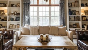 A cozy living room with a wooden ceiling, a large window with curtains, a sofa with cushions, a coffee table with candles, and shelves with framed pictures and books. - Historic Midtown Chateau + Guest House - Minutes from Everything (Memphis)