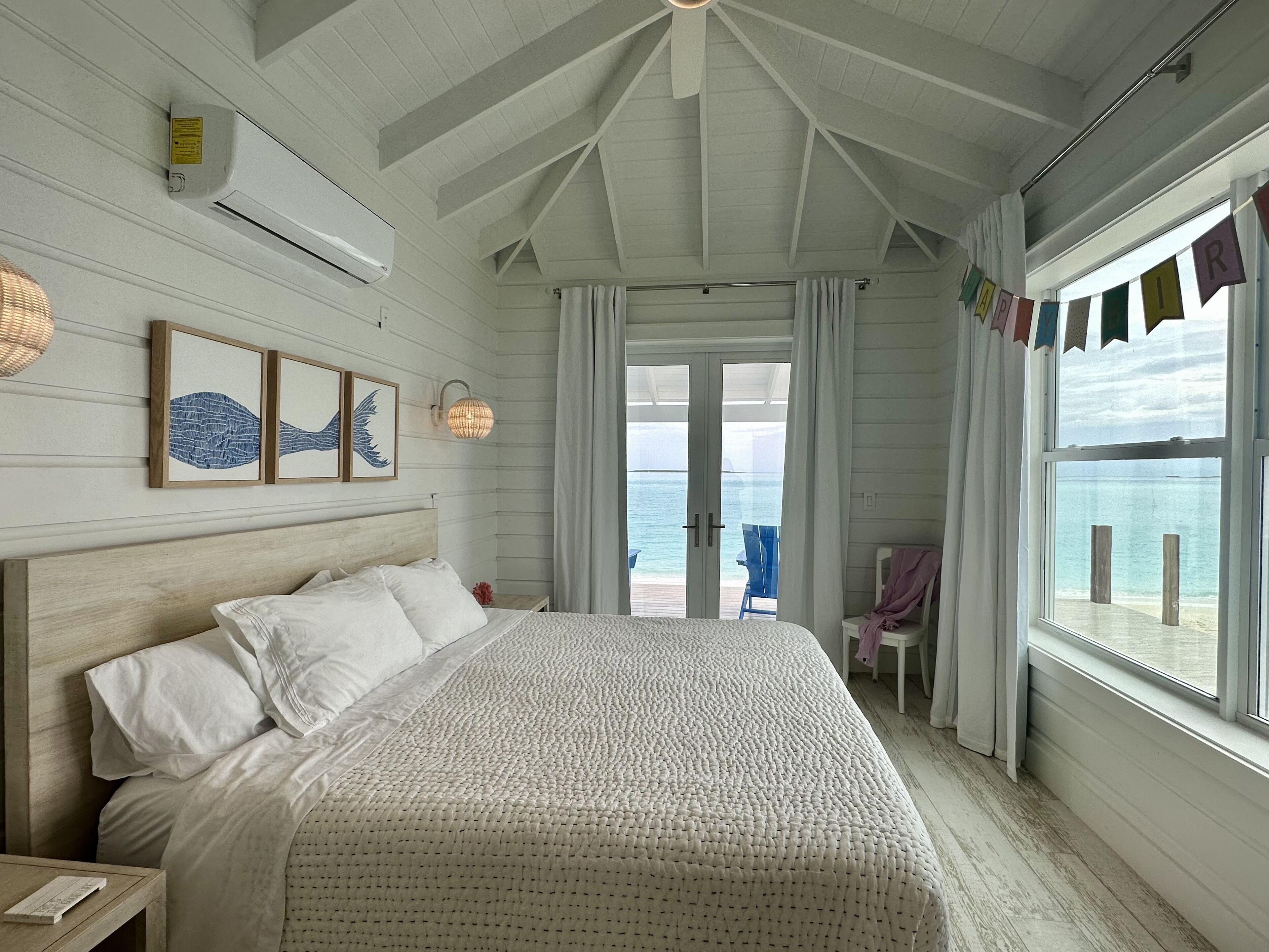 A bedroom with a large bed, a view of the sea through a glass door, and a ceiling fan.