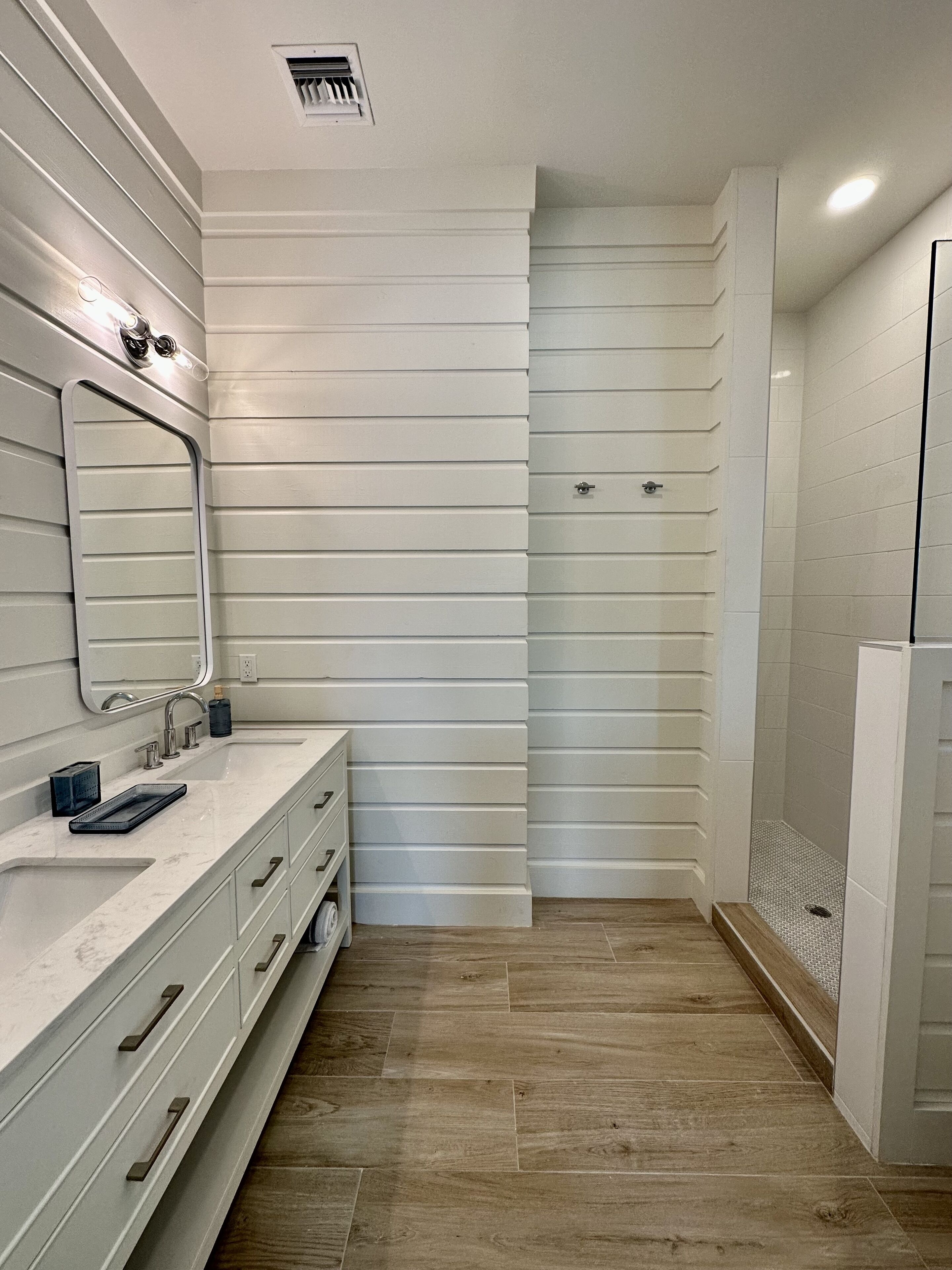A modern bathroom with a large mirror, two sinks, a shower, and wooden flooring.
