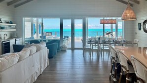 A spacious kitchen with a view of the ocean, a dining area with wicker chairs, and a large sliding glass door leading to a patio. - Luxury beachfront home, Heated private pool, Five King suites w/ ensuite baths! (Georgetown)
