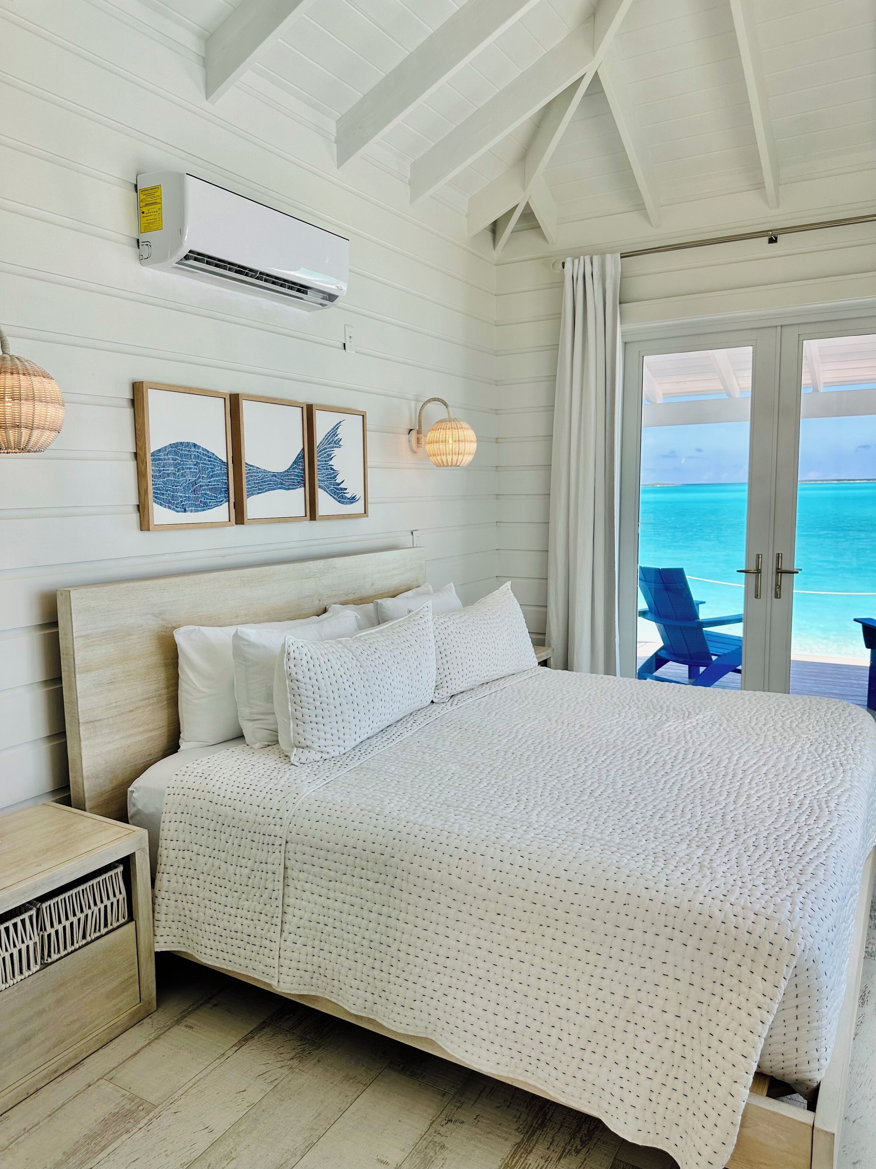 Une chambre avec un grand lit, du linge de lit blanc, une tête de lit en bois et une vue sur l’océan par une porte vitrée.