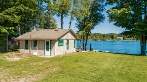 Une petite cabane en bois, dotée d’une porte et de fenêtres, située au bord d’un lac, avec une zone herbeuse devant.