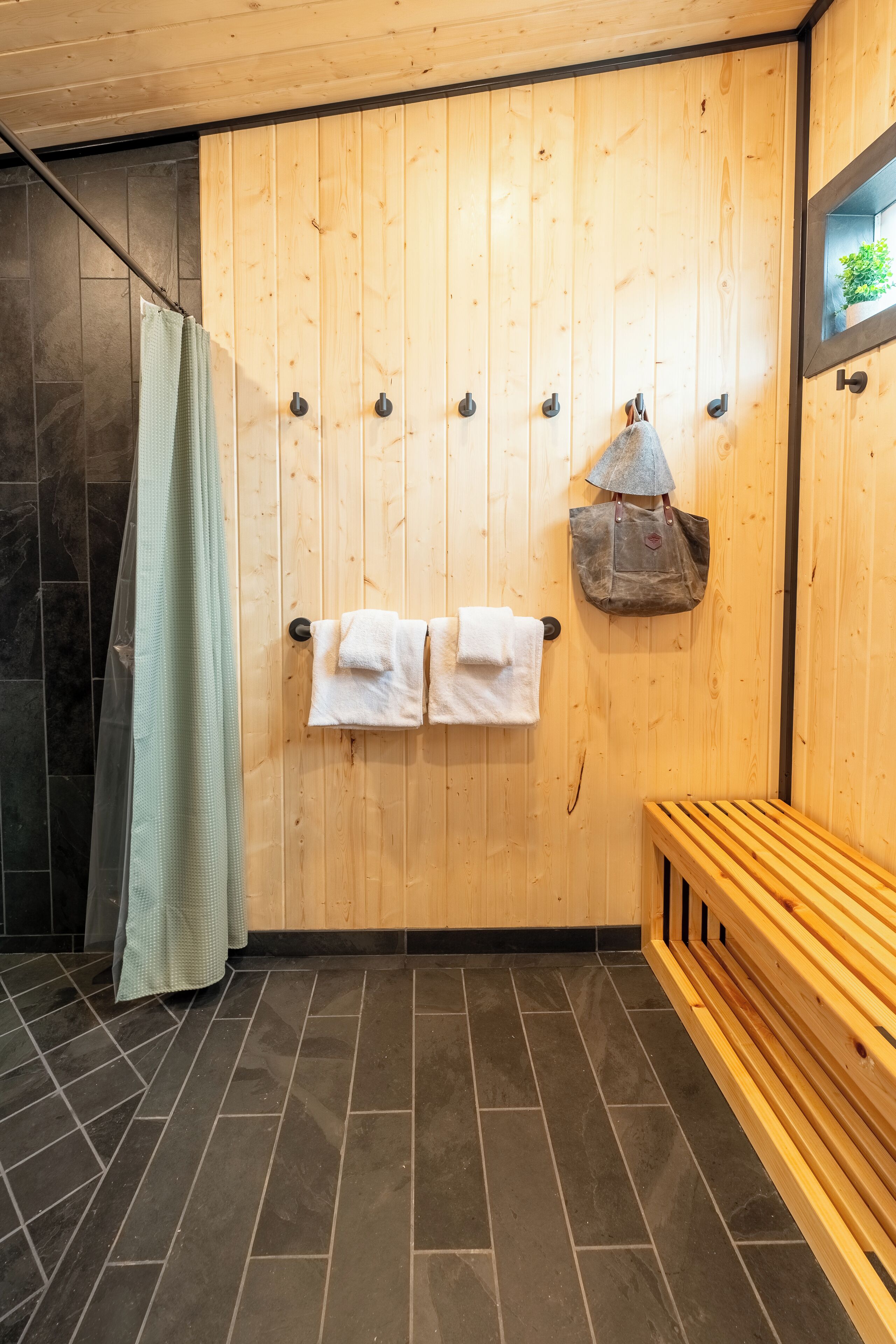 Um quarto com painéis de madeira, um banco, porta-toalhas com toalhas, uma bolsa pendurada e um cortinado de chuveiro.