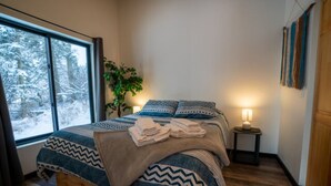 Ein Schlafzimmer mit großem Fenster, einem Bett mit gemusterten Betttextilien, einem Nachttisch mit Lampe und einer Pflanze.