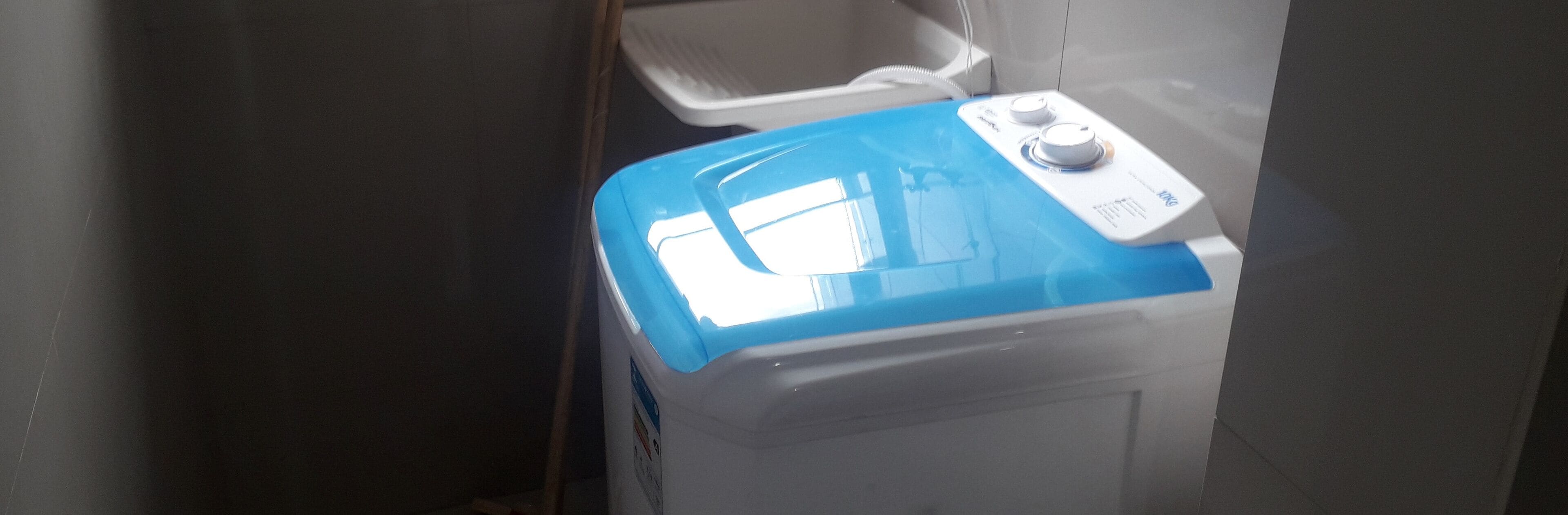 Una lavadora compacta con tapa azul, un panel de control y un recipiente de plástico blanco al fondo.