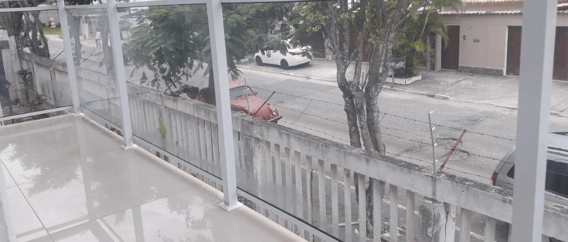 Ein Balkon mit Blick auf eine Straße, ein weißes Auto parkt am Straßenrand und ein Gebäude mit rotem Dach.