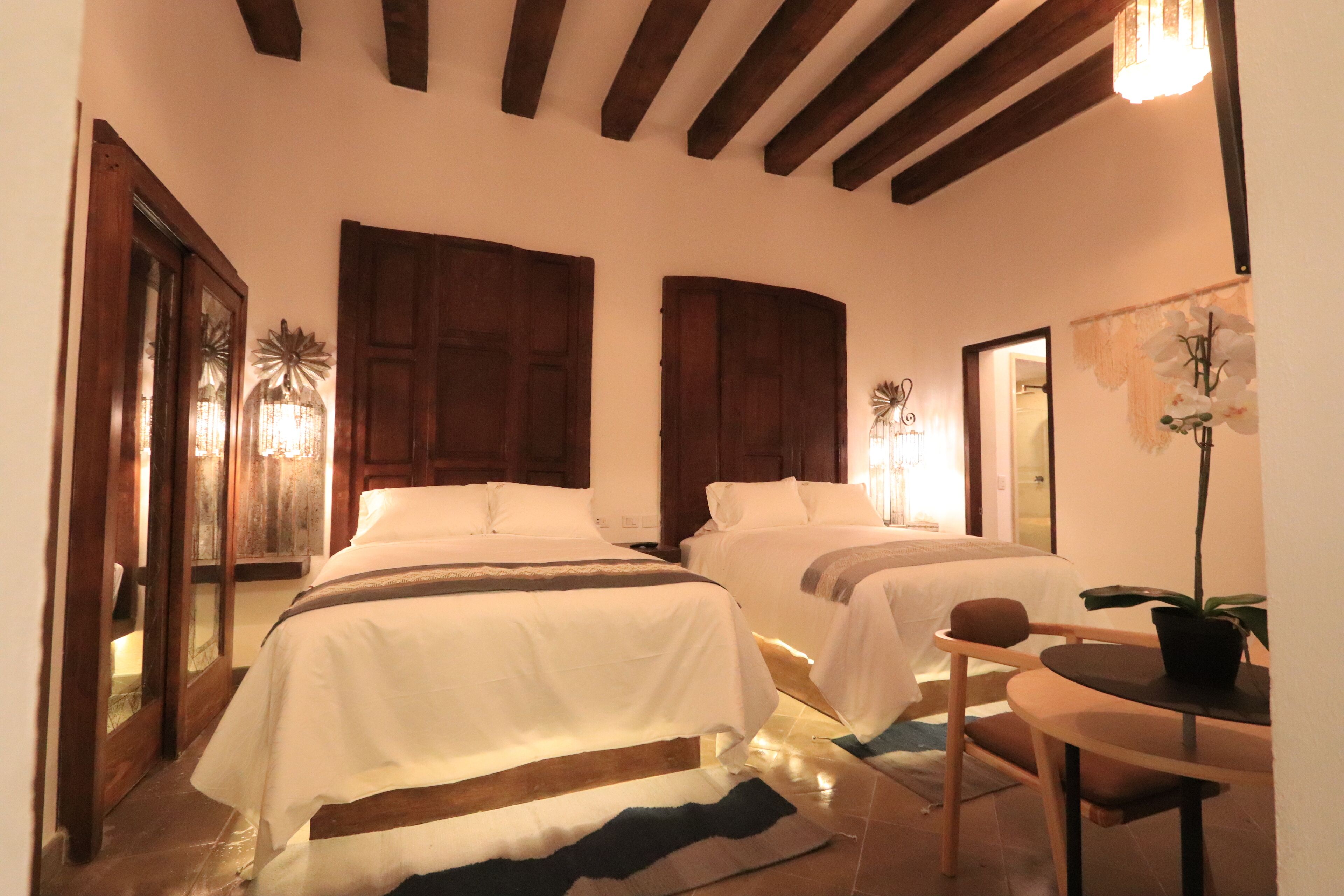 A hotel room with two beds, wooden beams on the ceiling, a small table with a chair, and a potted plant.