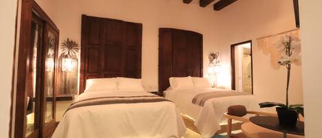A hotel room with two beds, wooden beams on the ceiling, a small table with a chair, and a potted plant.
