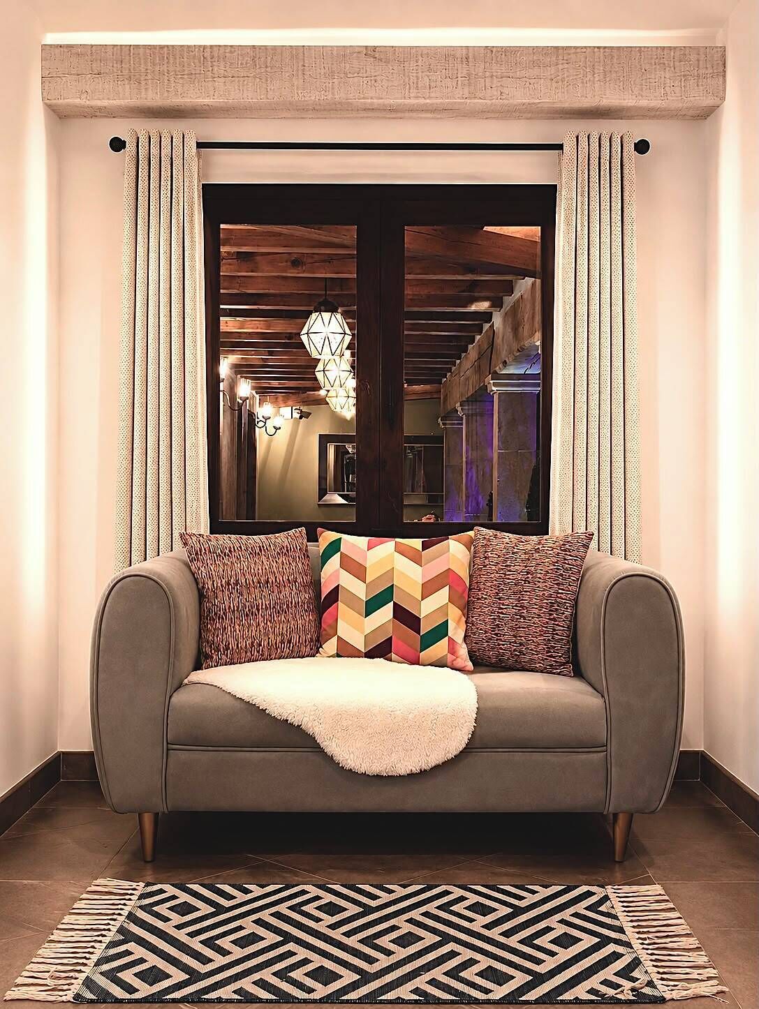 Un canapé agrémenté de coussins à motifs et recouvert d’un plaid, placé devant une fenêtre ornée de rideaux, le tout adossé à un mur dont le revêtement présente une texture particulière.