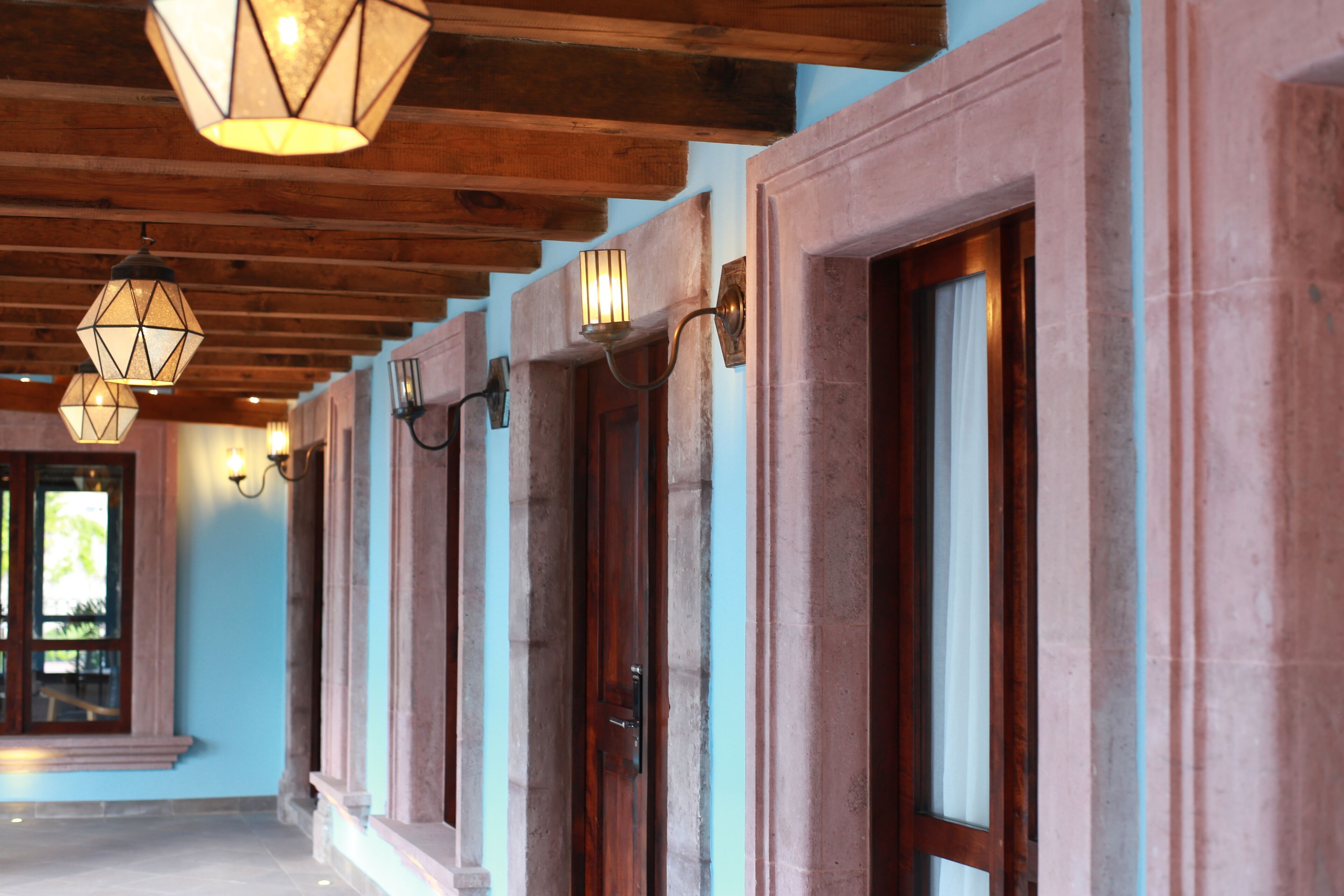 A hallway with wooden beams and stone columns, featuring doors with light fixtures above them.