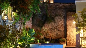 Una piscina con sillas de descanso, una pared de piedra y una torre de campanas de noche.