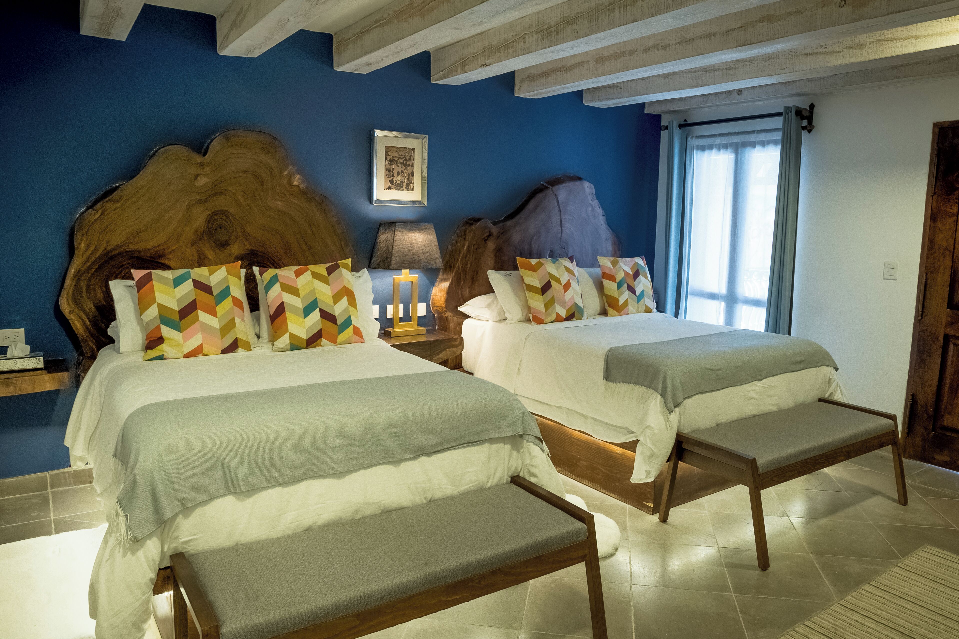 A hotel room with two beds, a wooden headboard, a nightstand with a lamp, and a large window with curtains.