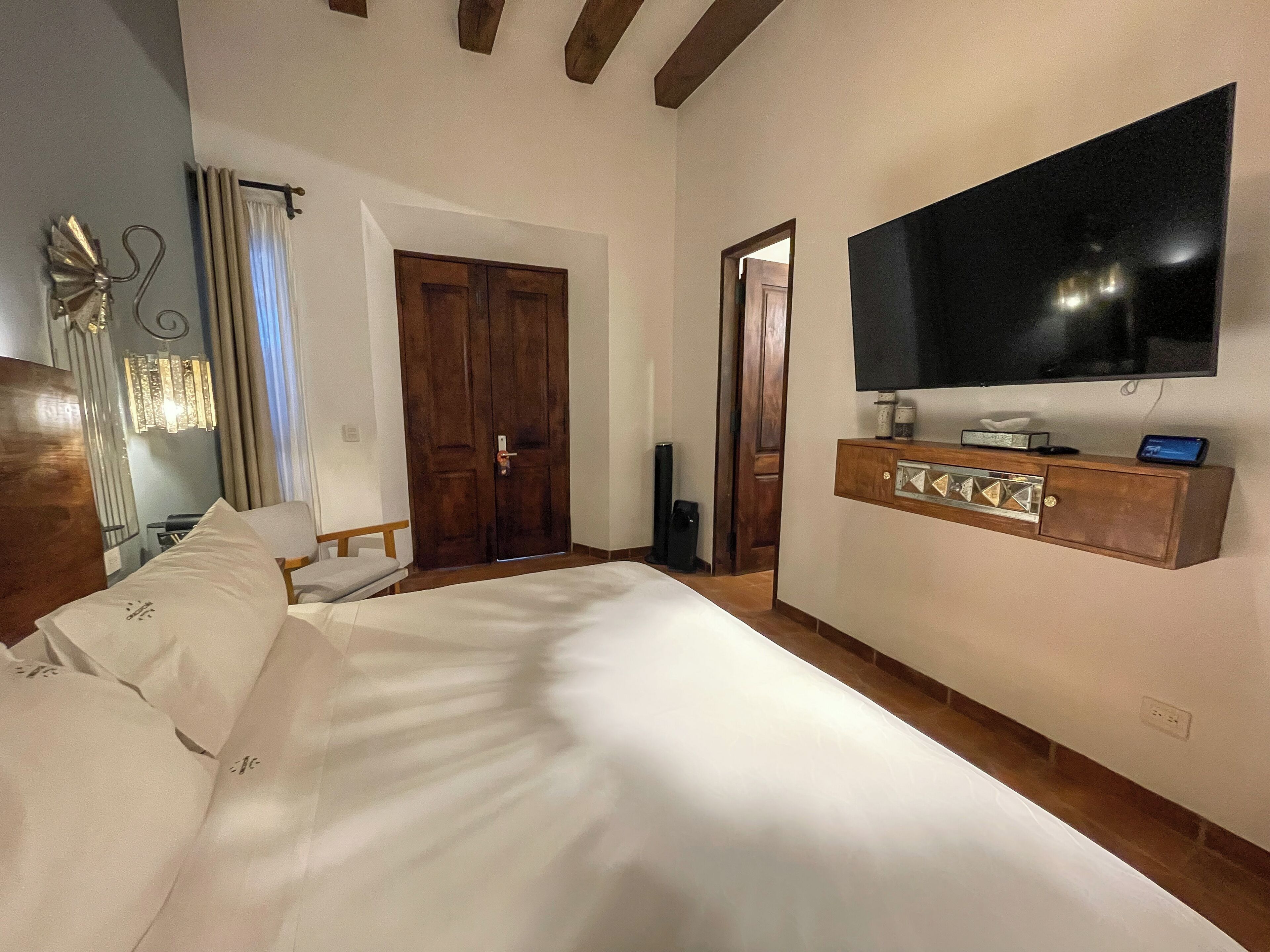 Hotelkamer met een groot bed, een aan de muur bevestigde televisie, een houten kastje onder de televisie en een deur die naar een andere kamer leidt.