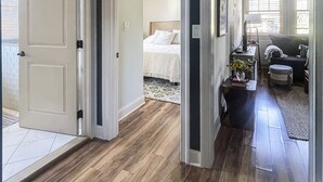 Un hall d’entrée spacieux, doté d’un plancher en bois franc, mène à une chambre à coucher équipée d’un lit et d’un ventilateur de plafond.