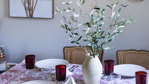 Une table à manger dressée avec des assiettes, des couverts et de la verrerie rouge, agrémentée d’un vase rempli de verdure et d’une estampe botanique encadrée accrochée au mur.