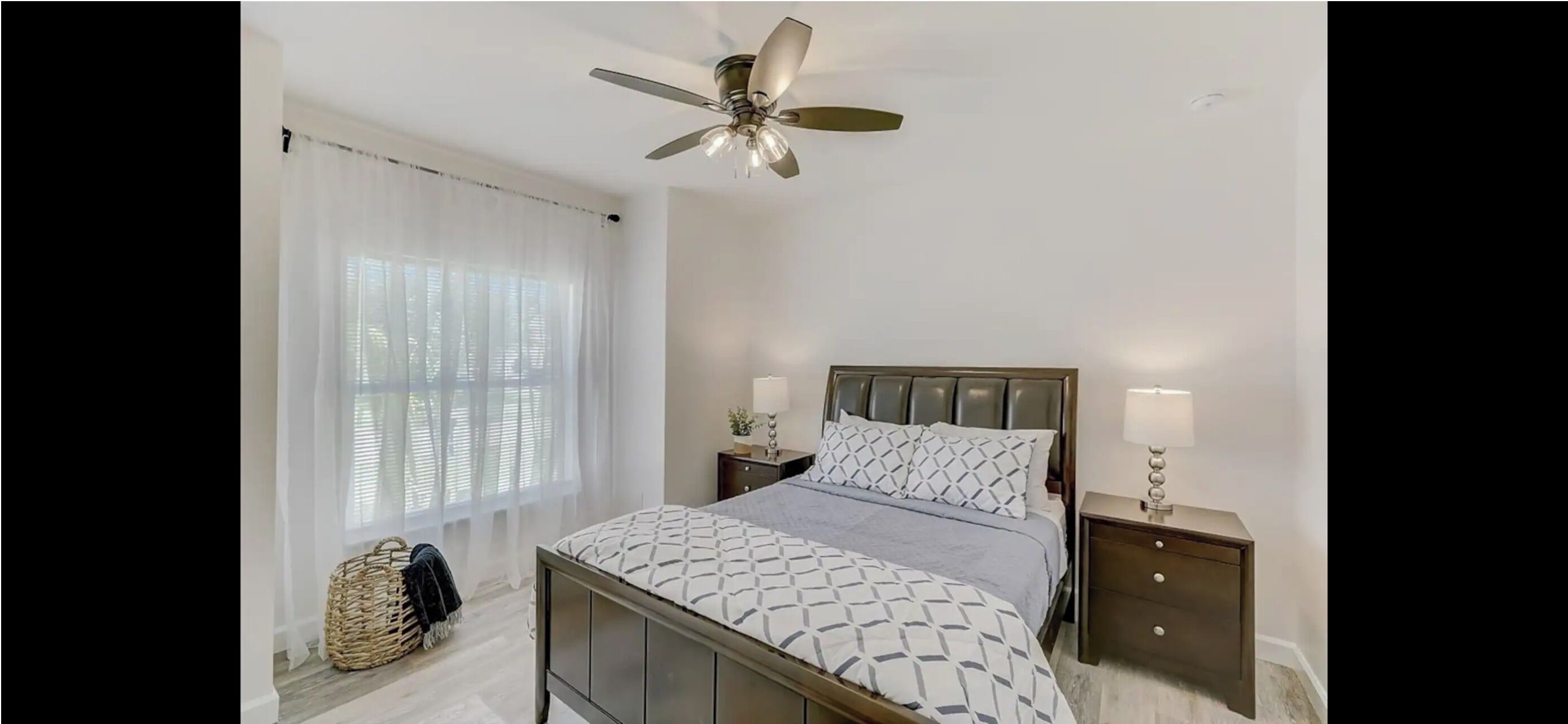 Une chambre à coucher avec un lit, un ventilateur de plafond, une table de chevet et une fenêtre avec des rideaux légers.