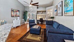 A living room with a large blue sofa, a small ottoman, a ceiling fan, and a dining area with a white table and chairs.