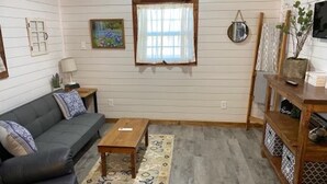 Una habitación con un sofá gris, una mesa de centro de madera, un tapete estampado, una ventana con cortina y un aire acondicionado de pared.