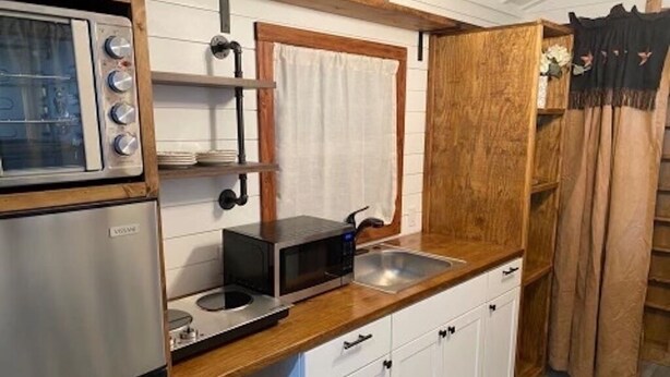 Cocina compacta con microondas, estufa y lavabo. Cuenta con gabinetes blancos y encimera de madera.