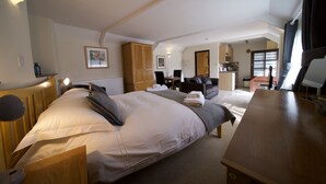 Een goed verlichte slaapkamer met een groot bed, een houten bureau, een stoel, een kledingkast en een kitchenette op de achtergrond.